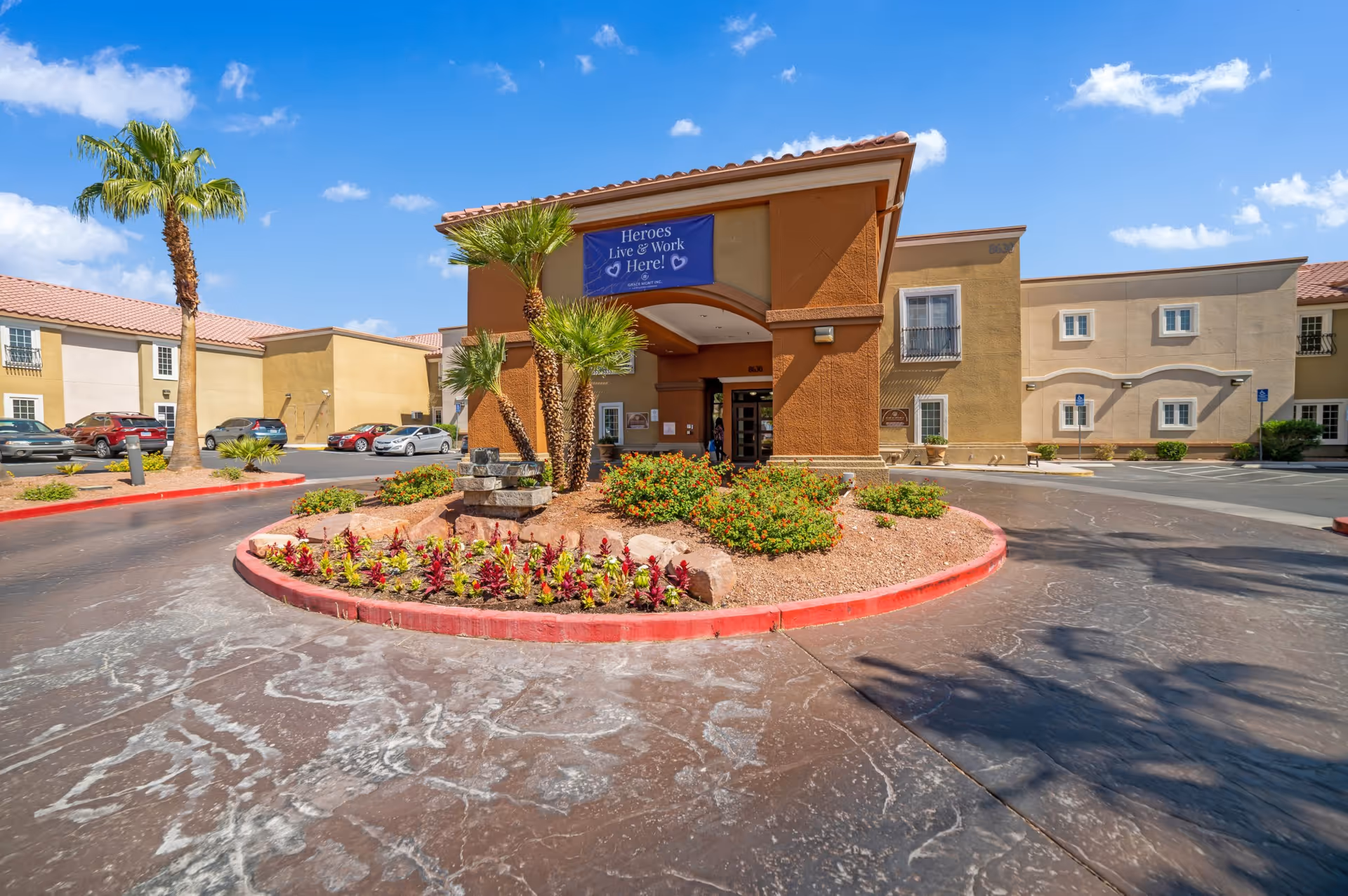 Exterior view of Acacia Springs facility entrance with a circular driveway, landscaped garden with palm trees and flowers, and a banner that reads 'Heroes Live & Work Here!' under a clear blue sky.
