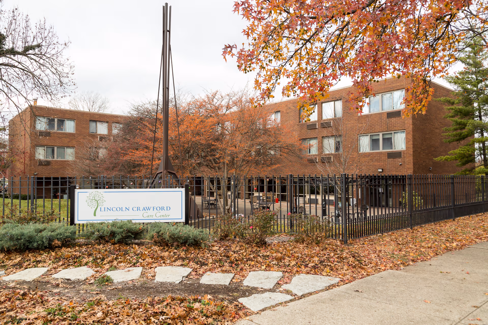 Front exterior of a brick senior care building with a fenced courtyard and a sign reading "Lincoln Crawford Care Center" amid autumn foliage.
