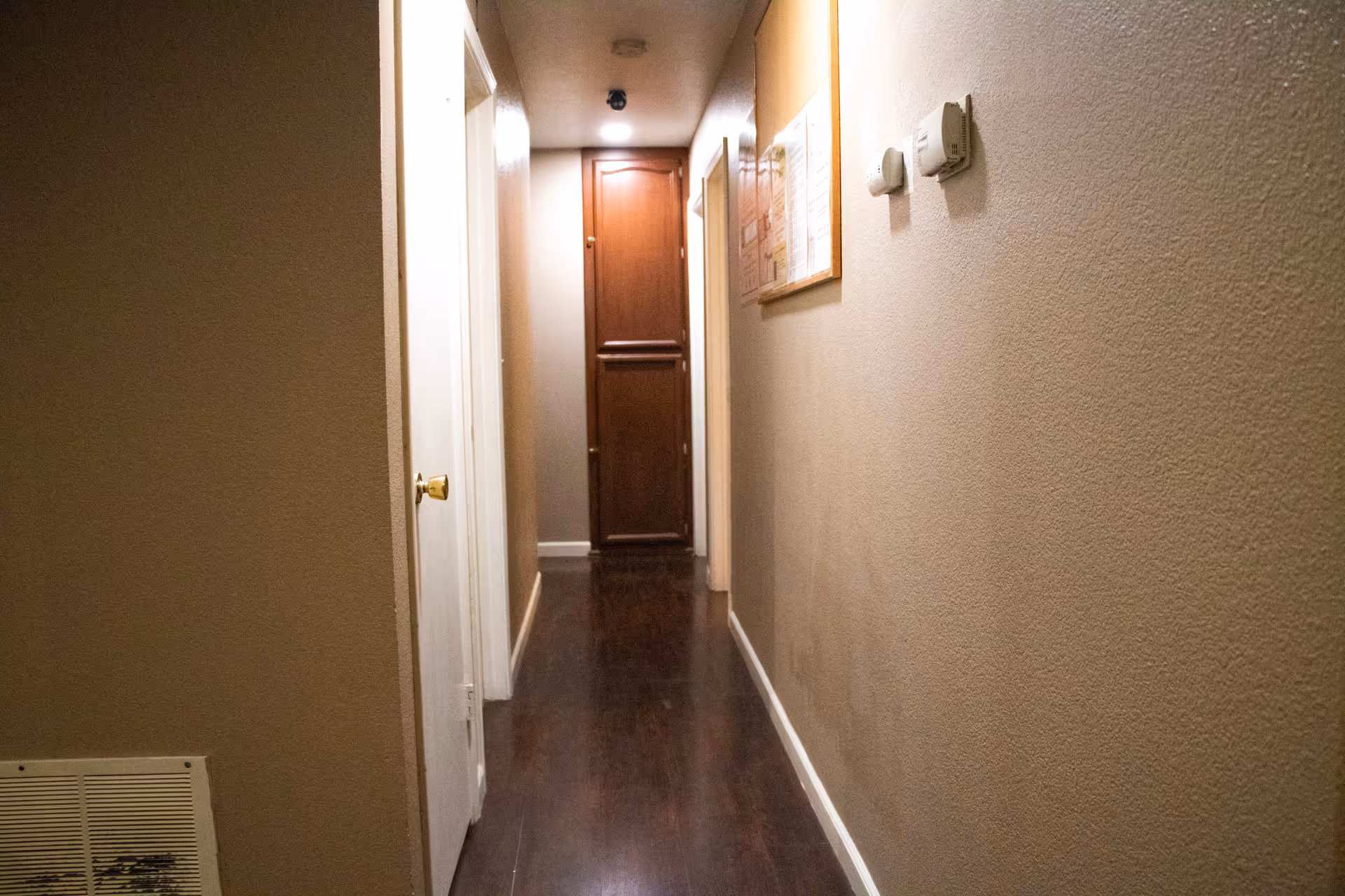 Long residential hallway with wood flooring, doors on both sides, and a bulletin board.