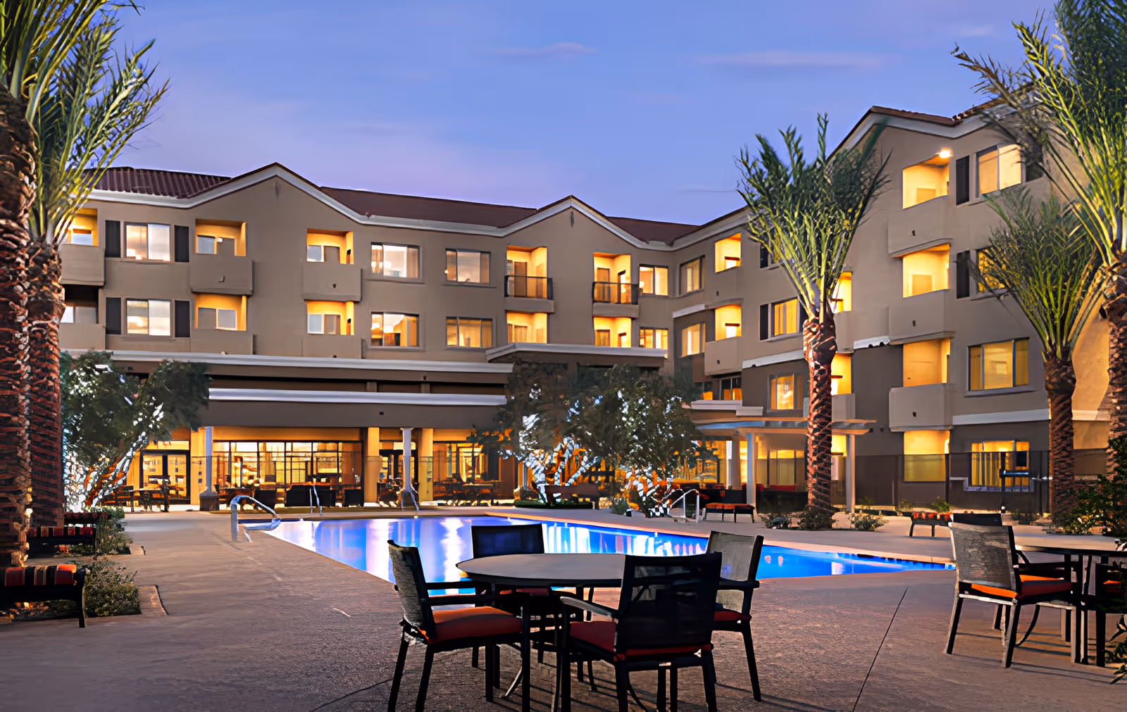 Dusk view of a multi-story residential building surrounding a lit outdoor swimming pool with patio tables and palm trees.