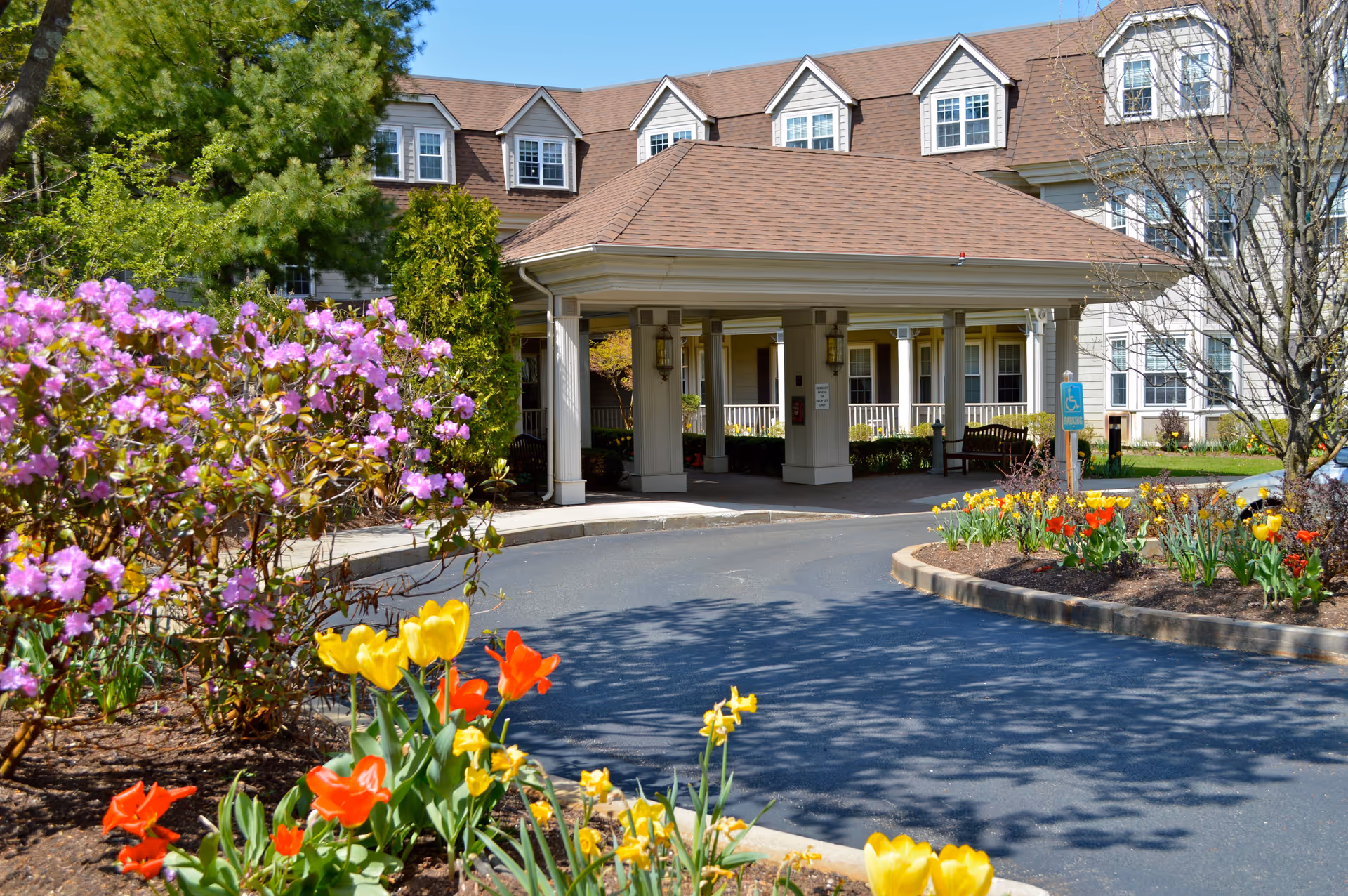 Entrance of a senior living facility with a covered drop-off area, surrounded by colorful flower beds including yellow and red tulips and purple flowering bushes, with a multi-story building in the background.