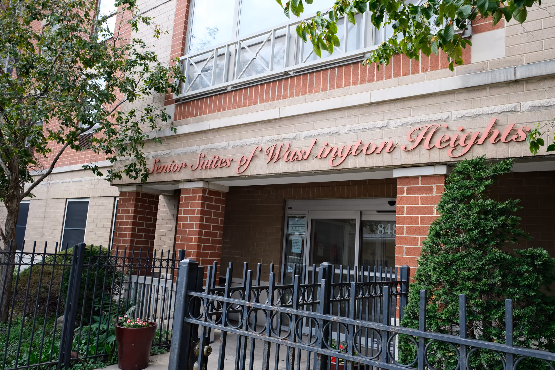 Entrance to Senior Suites of Washington Heights building with a black metal fence, potted plants, and greenery around the doorway. The building exterior is made of beige and red brick with a sign displaying the facility name above the entrance.