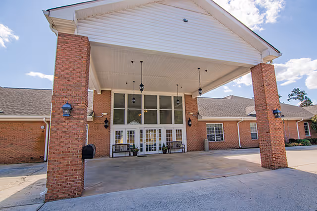 Front entrance of a brick building with a large covered driveway supported by brick columns. The entrance features glass double doors with sidelights and transom windows above. There are benches and potted plants near the doors, and outdoor wall lanterns mounted on the brick walls. The sky is partly cloudy.