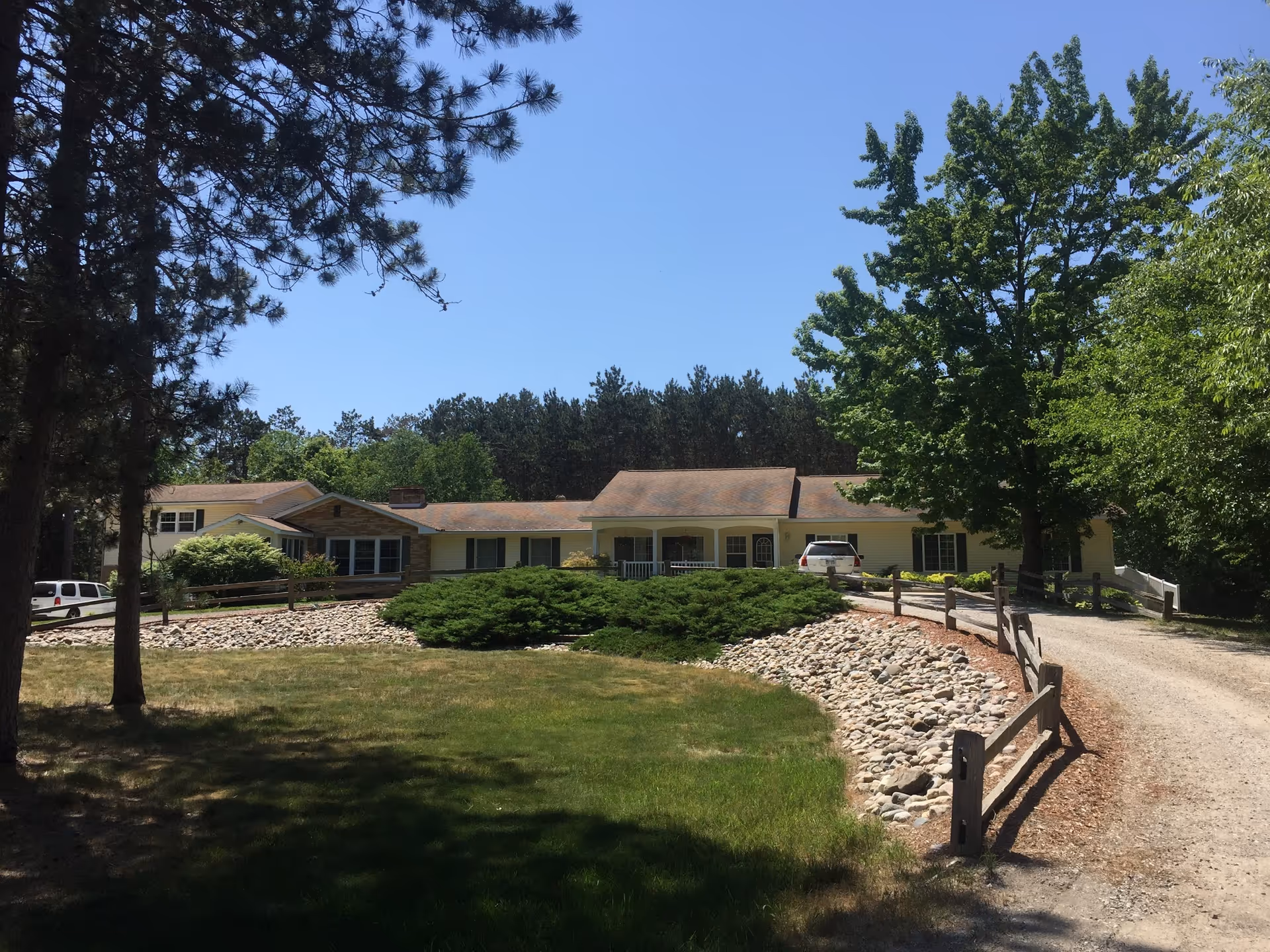 A single-story building with a light yellow exterior surrounded by trees and greenery under a clear blue sky. There is a gravel driveway leading up to the building, bordered by a wooden fence and landscaping with rocks and bushes.