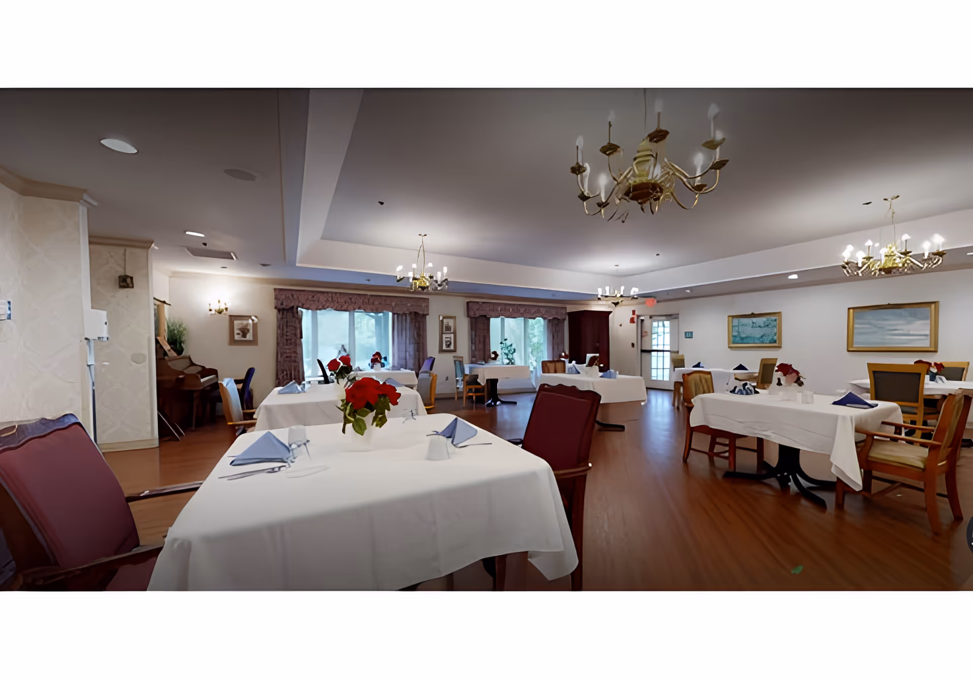 A bright dining room with tables set with white tablecloths, chairs, chandeliers, and floral centerpieces.
