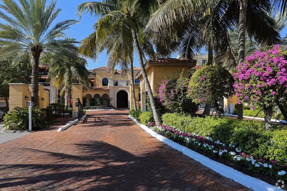 Entrance to a Mediterranean-style building with a brick driveway, palm trees and colorful landscaping.