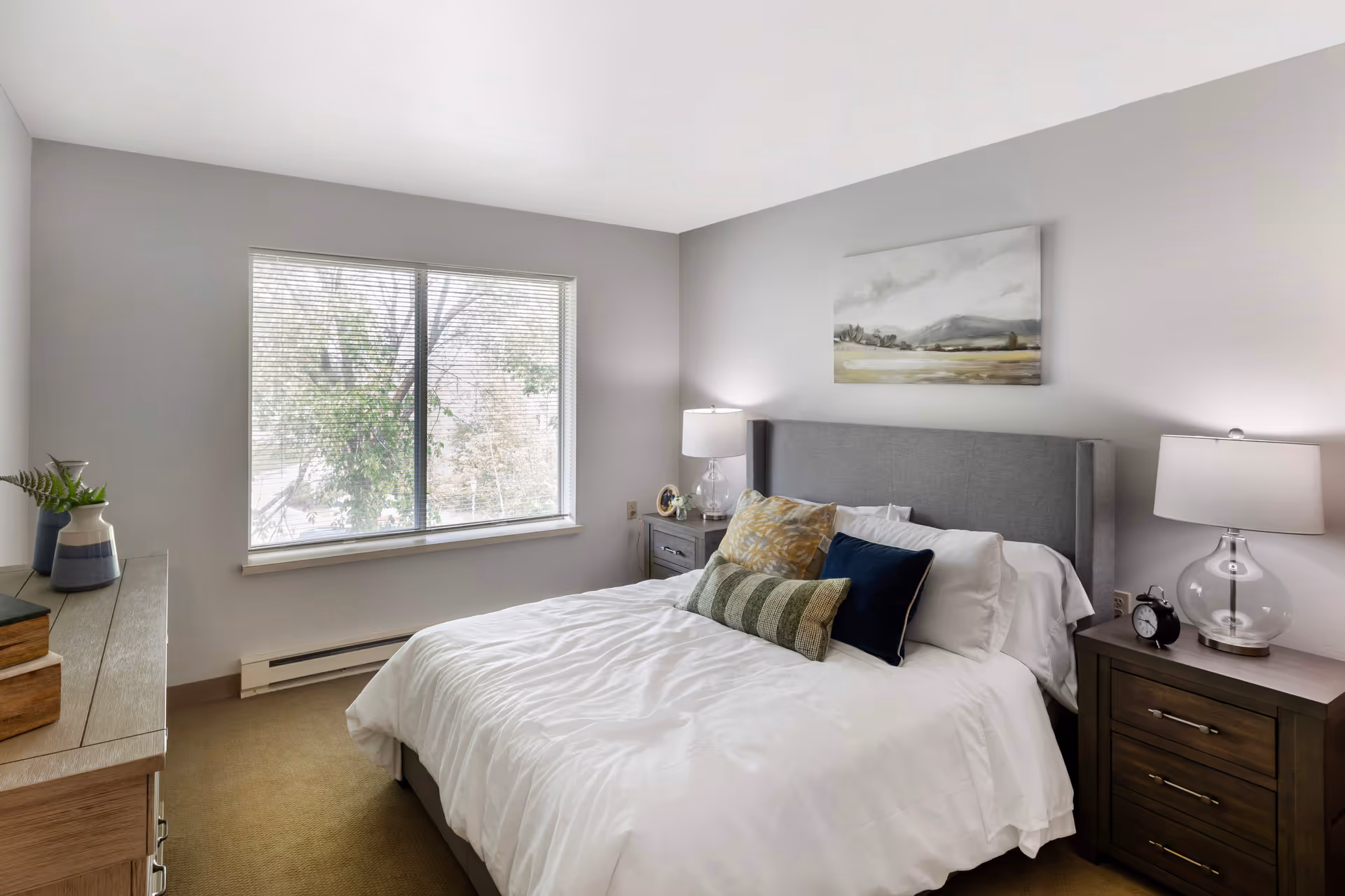 A neatly made bedroom with a grey upholstered headboard, two nightstands, lamps, and a window.