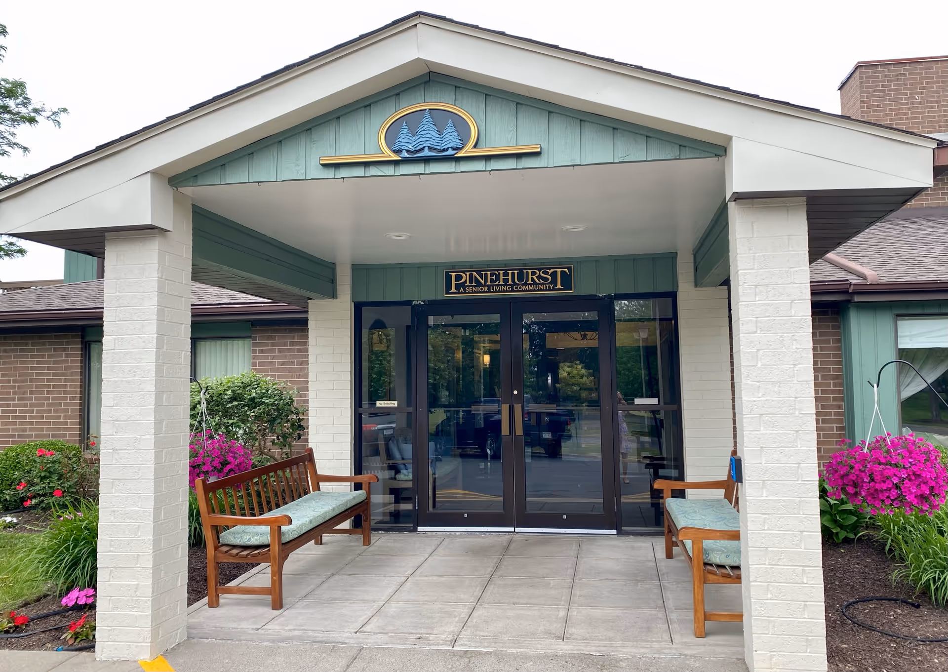 Front entry of Pinehurst Senior Living, with glass doors, benches, and colorful flowers.