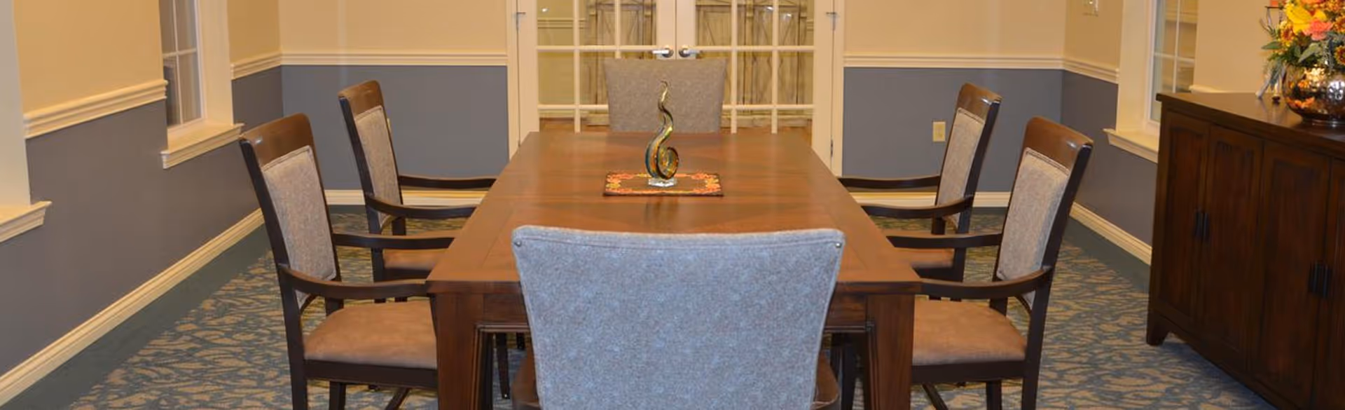 A formal dining room with a long wooden table surrounded by eight upholstered chairs. The room has blue and beige walls with white trim, carpeted floor with a patterned design, and a wooden sideboard with a decorative vase and flowers on the right side. Double doors with glass panels are visible at the far end of the room.