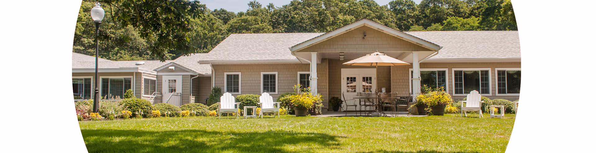 Exterior view of a single-story senior living facility building with beige siding, white trim, and multiple windows. In front of the building is a well-maintained lawn with several white Adirondack chairs, small tables, potted plants, and a patio area with a table and umbrella. Trees and greenery surround the building.