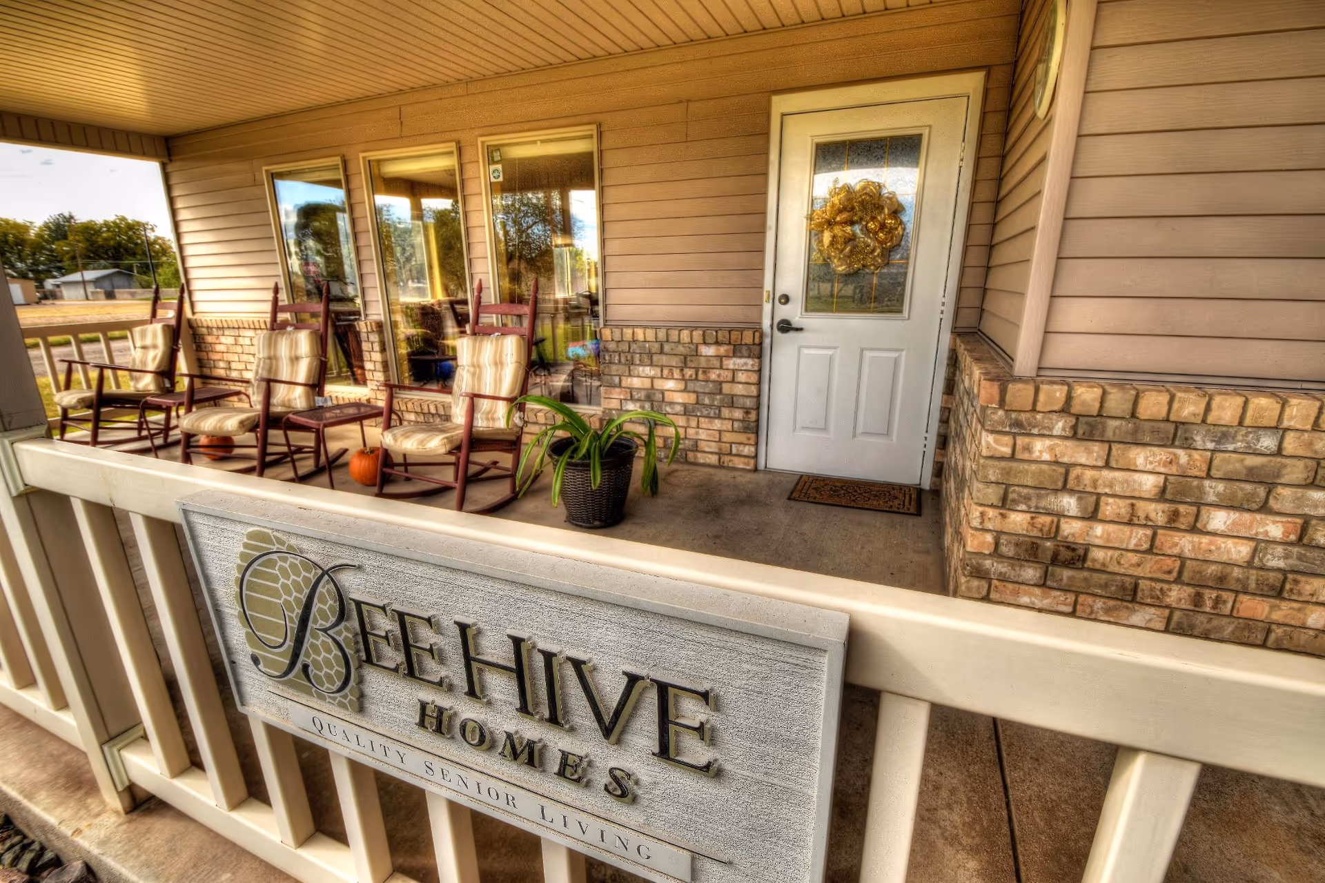 Front porch of BeeHive Homes of Portales featuring a white door with a decorative wreath, three cushioned rocking chairs, a potted plant, and a sign on the railing that reads 'BeeHive Homes Quality Senior Living'. The porch has beige siding and brick accents.