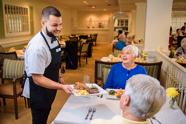 A smiling server brings plated meals to elderly residents seated at a dining table in a bright communal dining room.