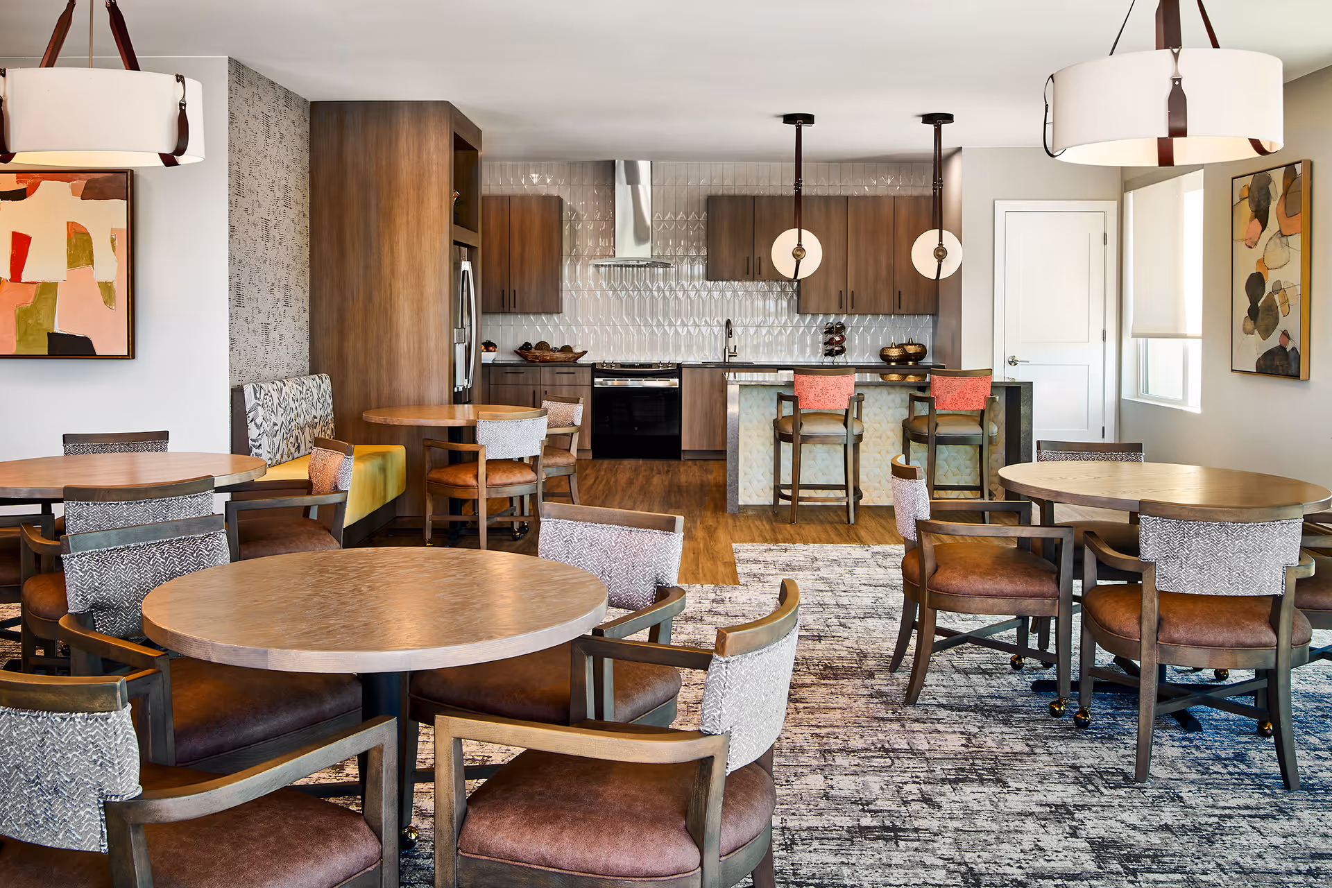 A modern dining area with multiple round wooden tables and cushioned chairs arranged around them. In the background, there is a kitchen with wooden cabinets, a stainless steel refrigerator, stove, and a kitchen island with two bar stools. The room is decorated with abstract paintings on the walls and two large pendant lights hanging from the ceiling. The floor has a combination of wood and patterned carpet.