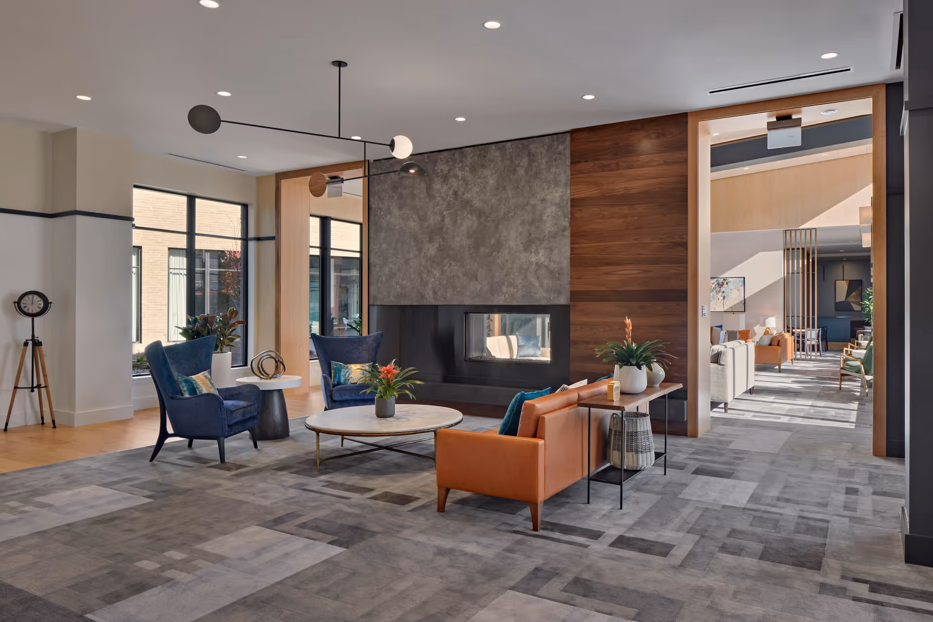 A modern and spacious living room area in a senior living facility featuring a large gray patterned carpet, two blue armchairs, a round marble coffee table with a plant, and a brown leather sofa with pillows. The room has a contemporary fireplace with a concrete and wood accent wall, large windows letting in natural light, and a stylish black and white ceiling light fixture. In the background, another seating area is visible through a large open doorway.