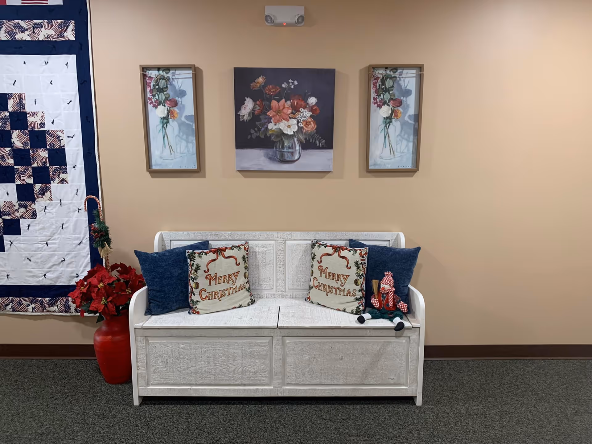 A white wooden bench with two blue pillows and two 'Merry Christmas' decorative pillows. A small Christmas doll sits on the right side of the bench. Above the bench are three framed floral paintings. To the left of the bench is a red vase with poinsettia flowers and a quilt hanging on the wall.