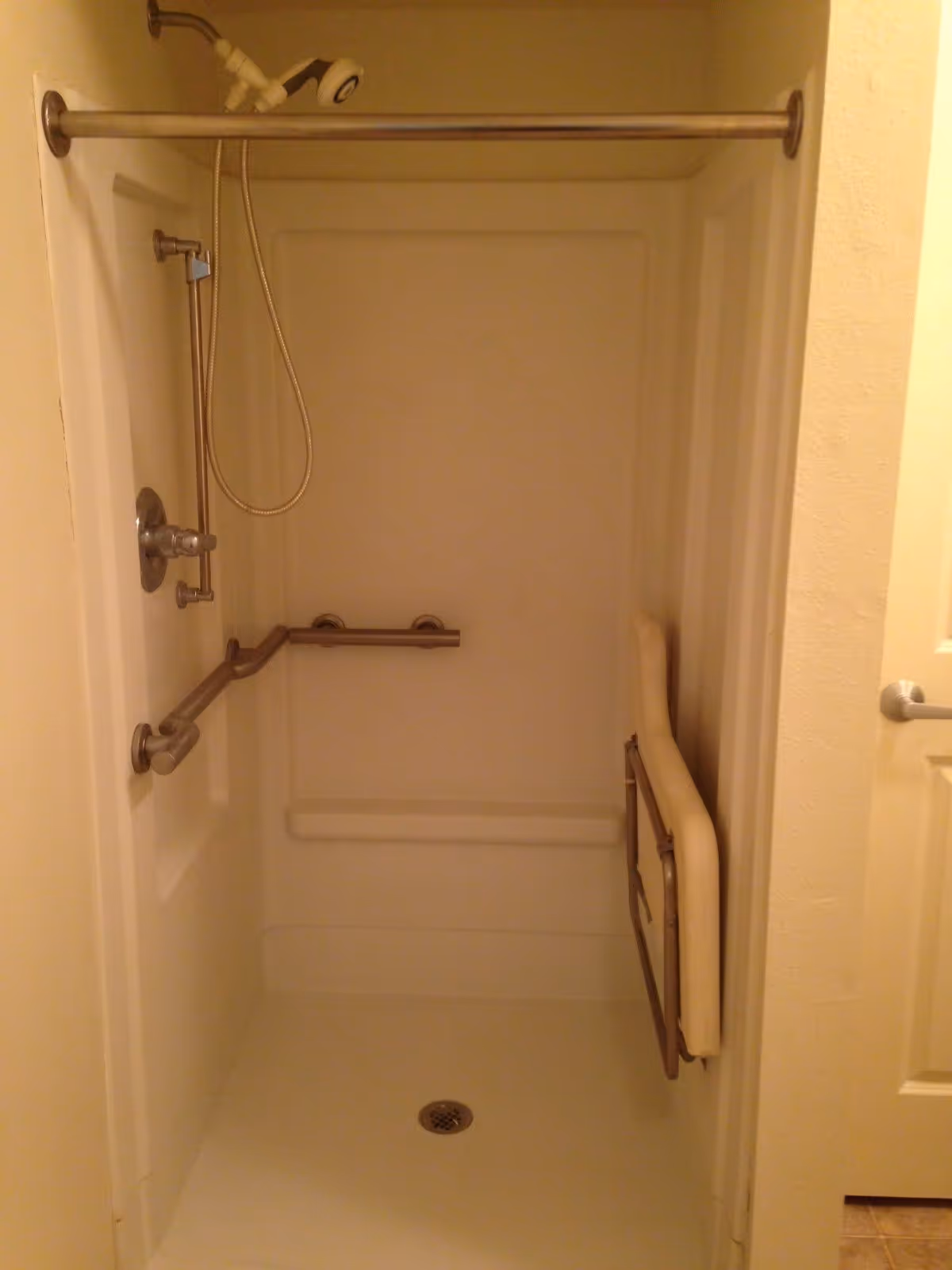A walk-in shower with grab bars on the walls and a fold-down seat on the right side. The shower has a handheld showerhead attached to a vertical sliding bar and a horizontal grab bar for support. The shower floor has a drain in the center.