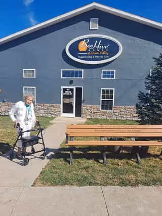An elderly woman using a walker is standing on a sidewalk in front of a building with a sign that reads 'BeeHive Homes of Grain Valley Assisted Living & Memory Care.' There is a wooden bench on the grass near the sidewalk, and the building has a gray exterior with stone accents and several windows.