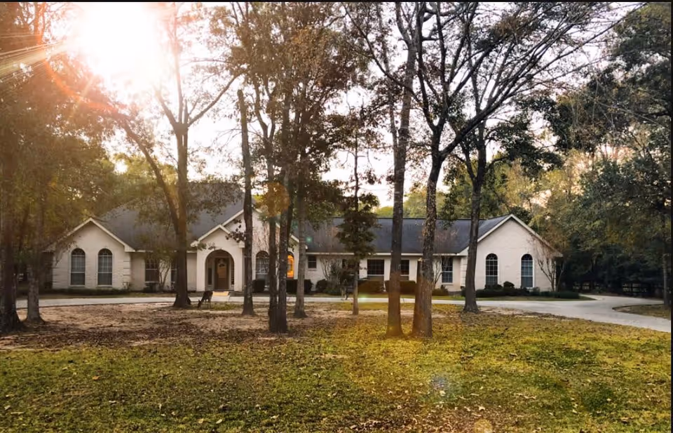 A single-story building with a light-colored exterior surrounded by tall trees and a grassy area in front. The sun is shining through the trees, creating a warm glow. A curved driveway leads to the entrance of the building.