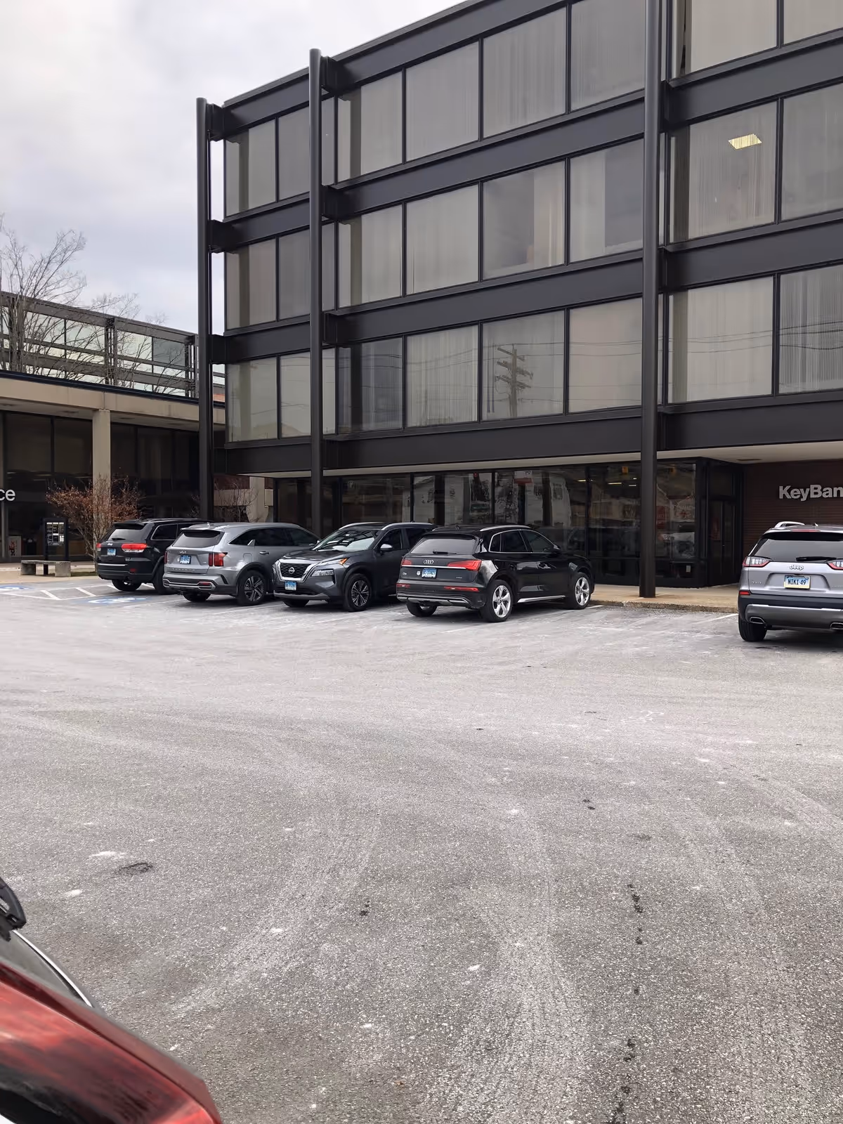 Modern glass-fronted building with several parked cars in a spacious parking lot.