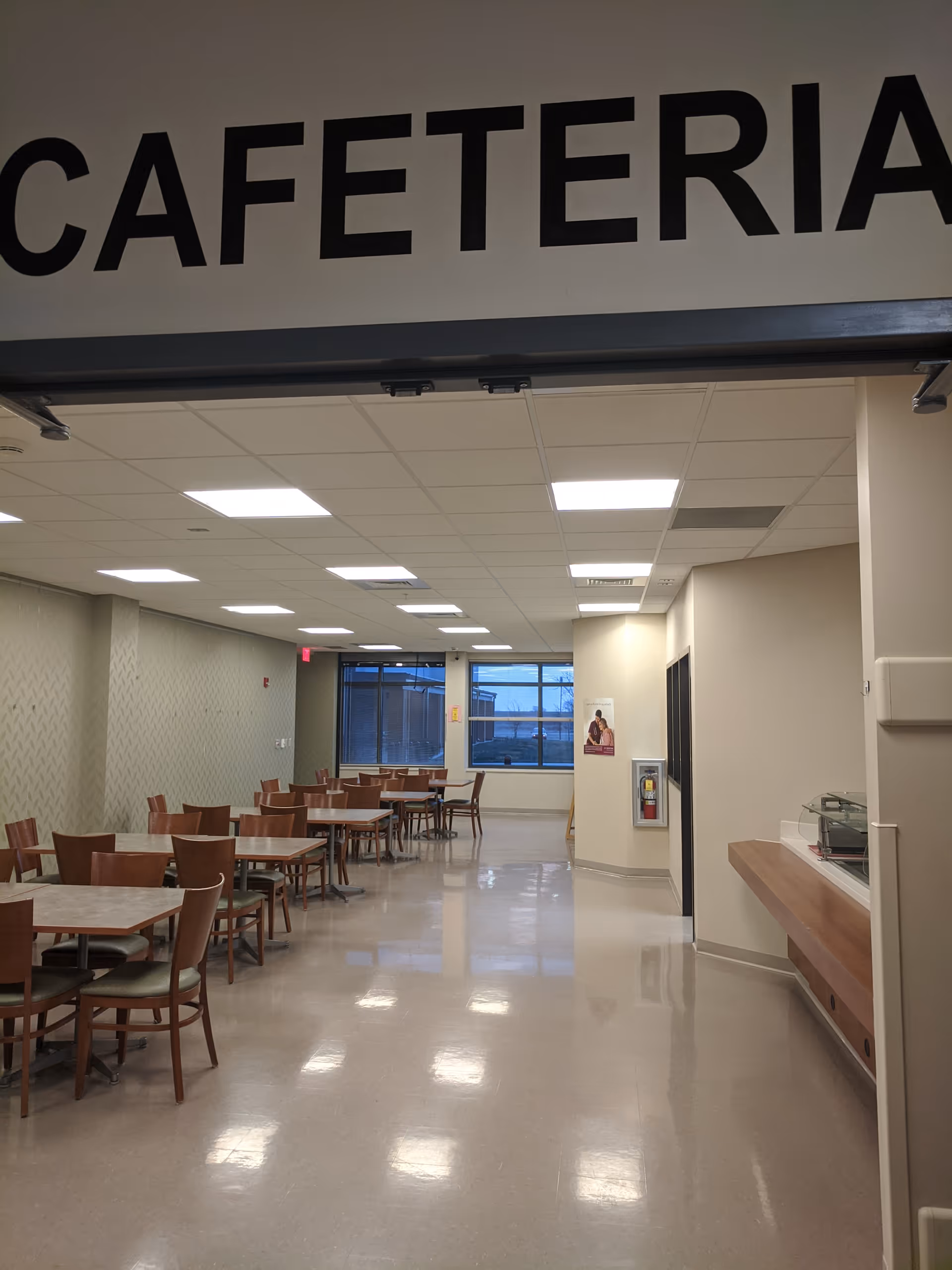 Long cafeteria with many tables and chairs, bright ceiling lights, and a serving counter.