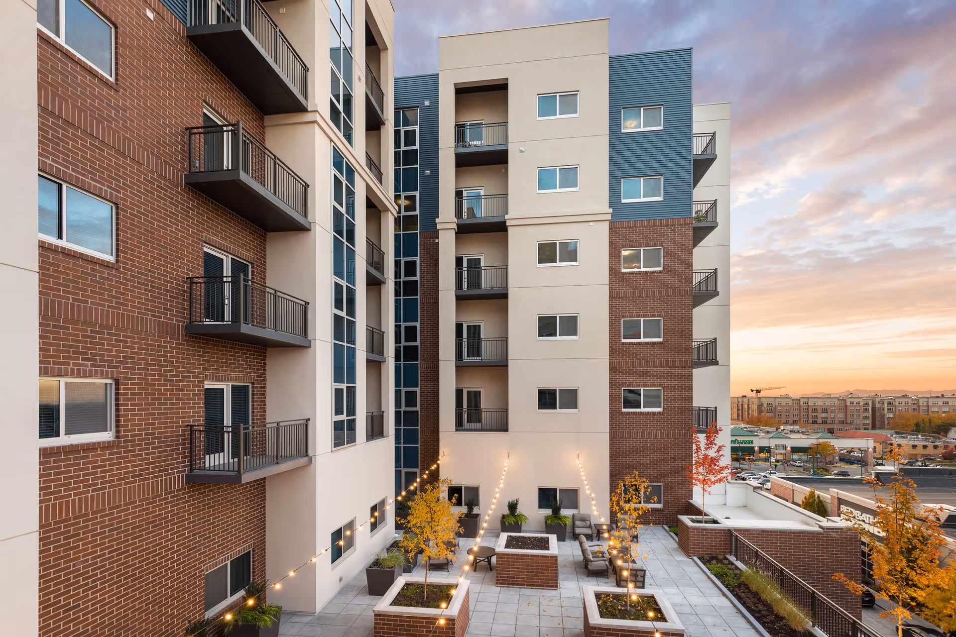 Outdoor courtyard area of a multi-story residential building with brick and beige exterior walls, small balconies, string lights, potted plants, and seating areas. The sky is partly cloudy with a sunset in the background.