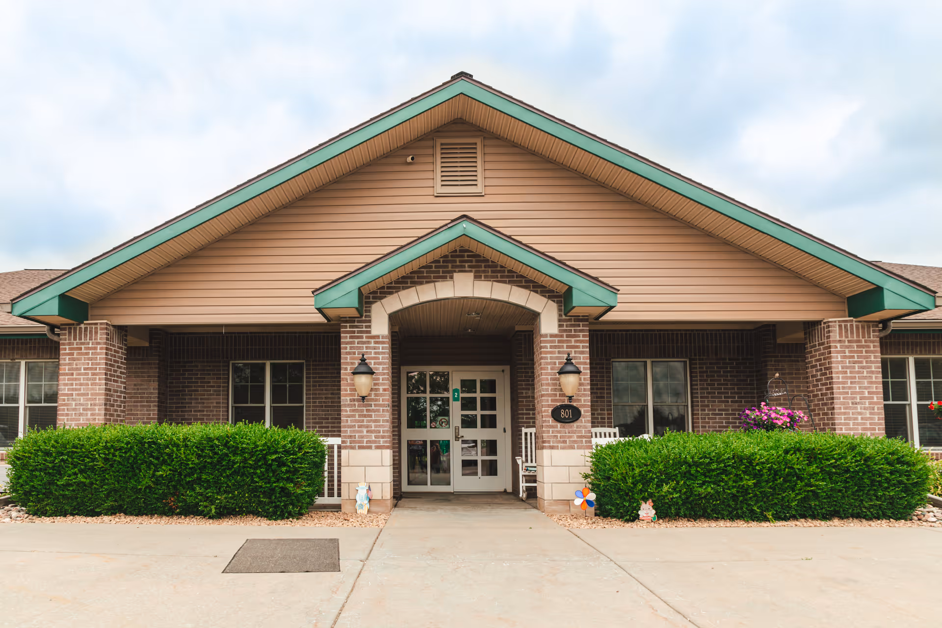 Front exterior view of a single-story brick building with a peaked roof and green trim. The entrance has double glass doors flanked by two wall-mounted lantern-style lights. There are neatly trimmed bushes on either side of the entrance and a concrete walkway leading up to the door. The building number 801 is displayed near the entrance.