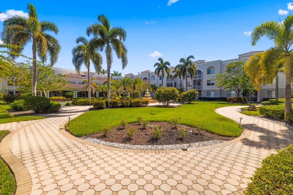 Sunny courtyard of a senior living facility with palm trees, curved walkways, and a white building.