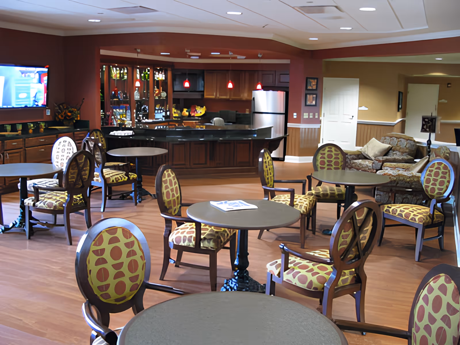 A spacious common area in Elmwood Assisted Living of New Bremen featuring several round tables with patterned chairs, a kitchen area with a refrigerator and hanging red pendant lights, a wall-mounted TV, and a comfortable seating area with sofas in the background.