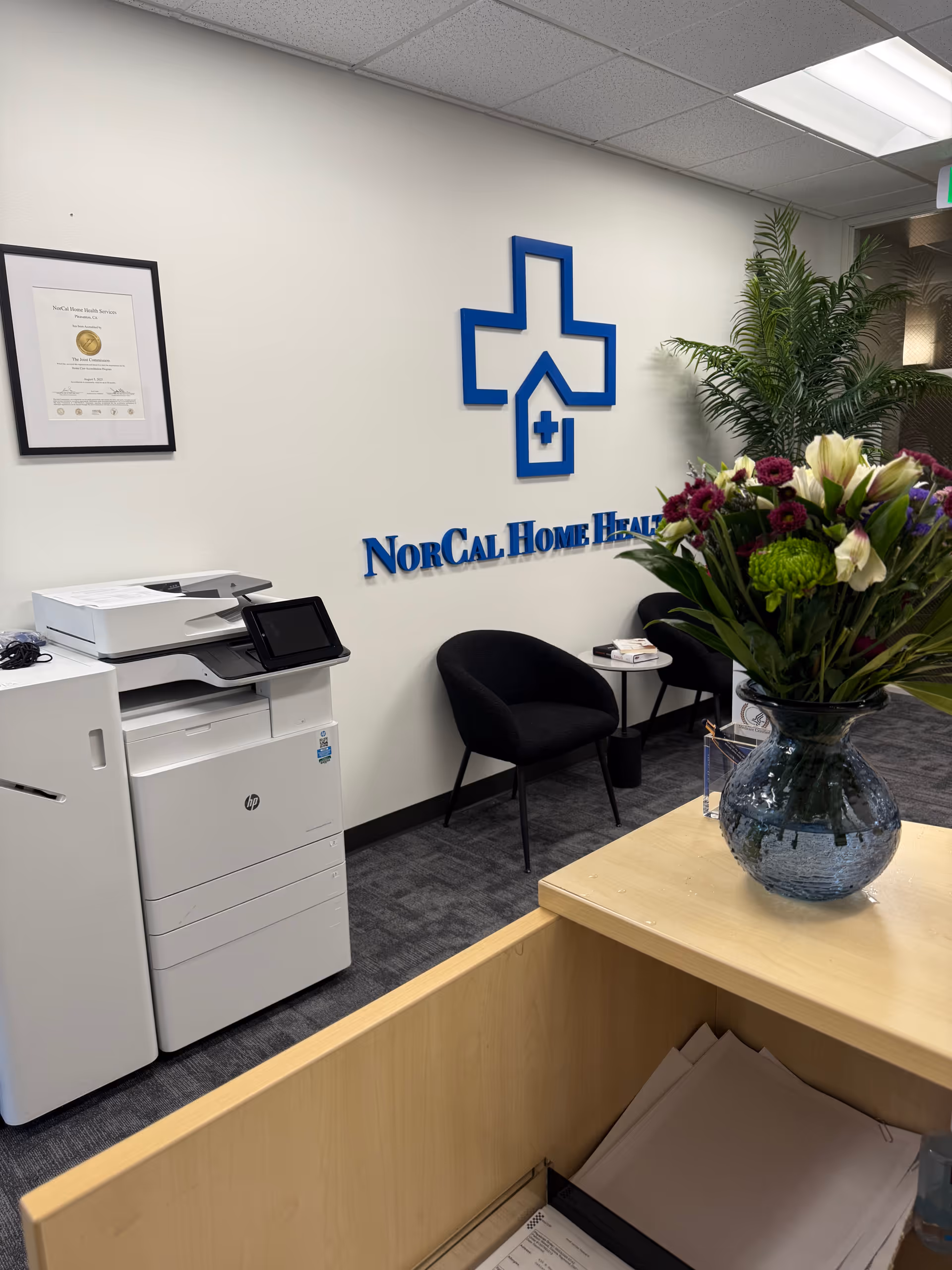 Reception area of NorCal Home Health Services featuring wall logo, flowers, chairs, and a printer.