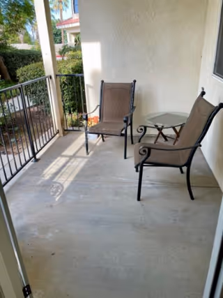 A covered outdoor patio area with two brown metal chairs and a small round glass-top table. The patio has a concrete floor and black metal railing, with greenery and part of a building visible in the background.