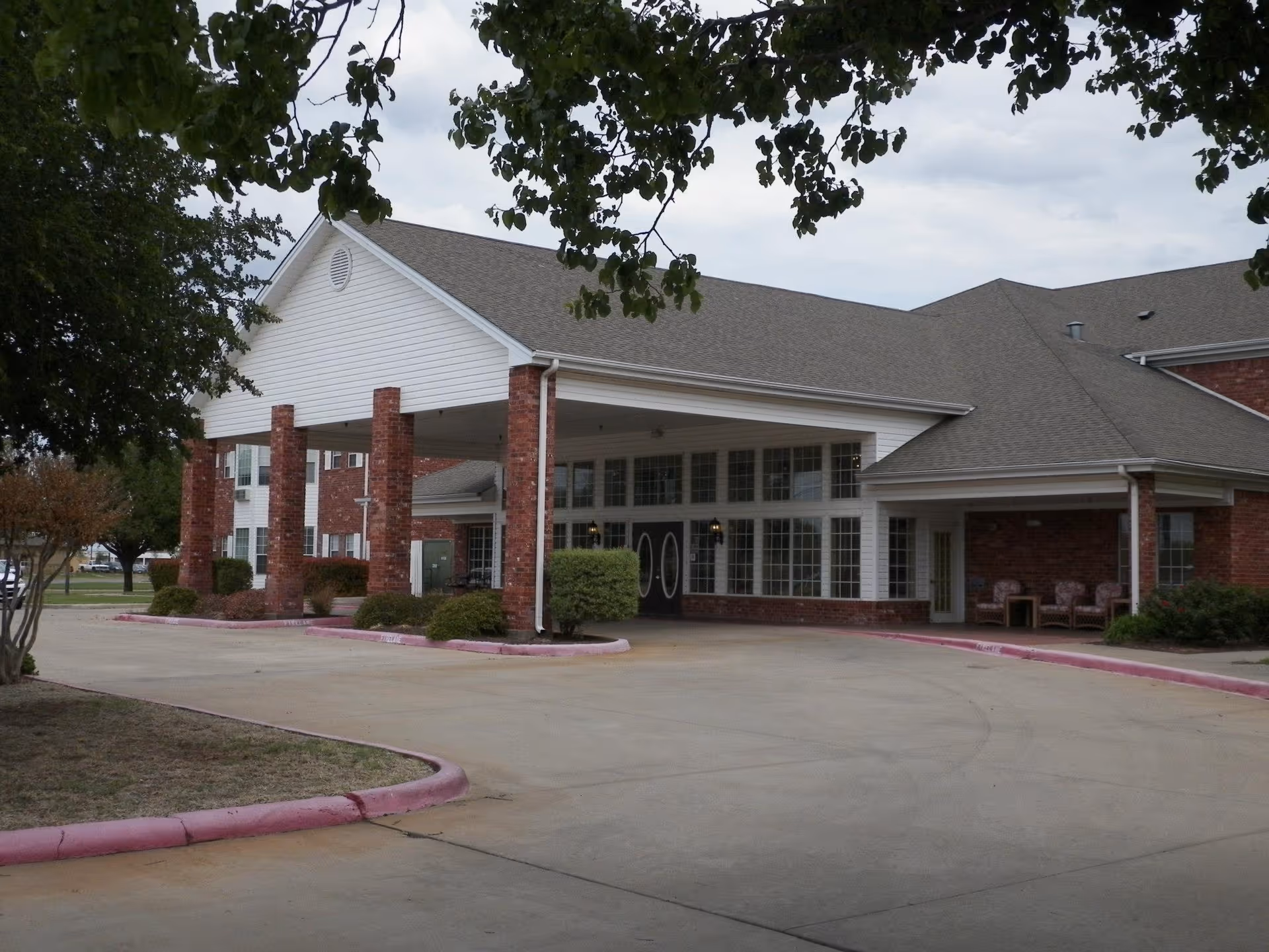 Front entrance of a brick senior living building with a covered porte-cochère, large windows, and surrounding landscaping.