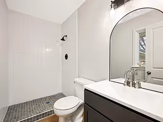 Modern bathroom with white tiles, shower area, toilet, and dark vanity with curved mirror