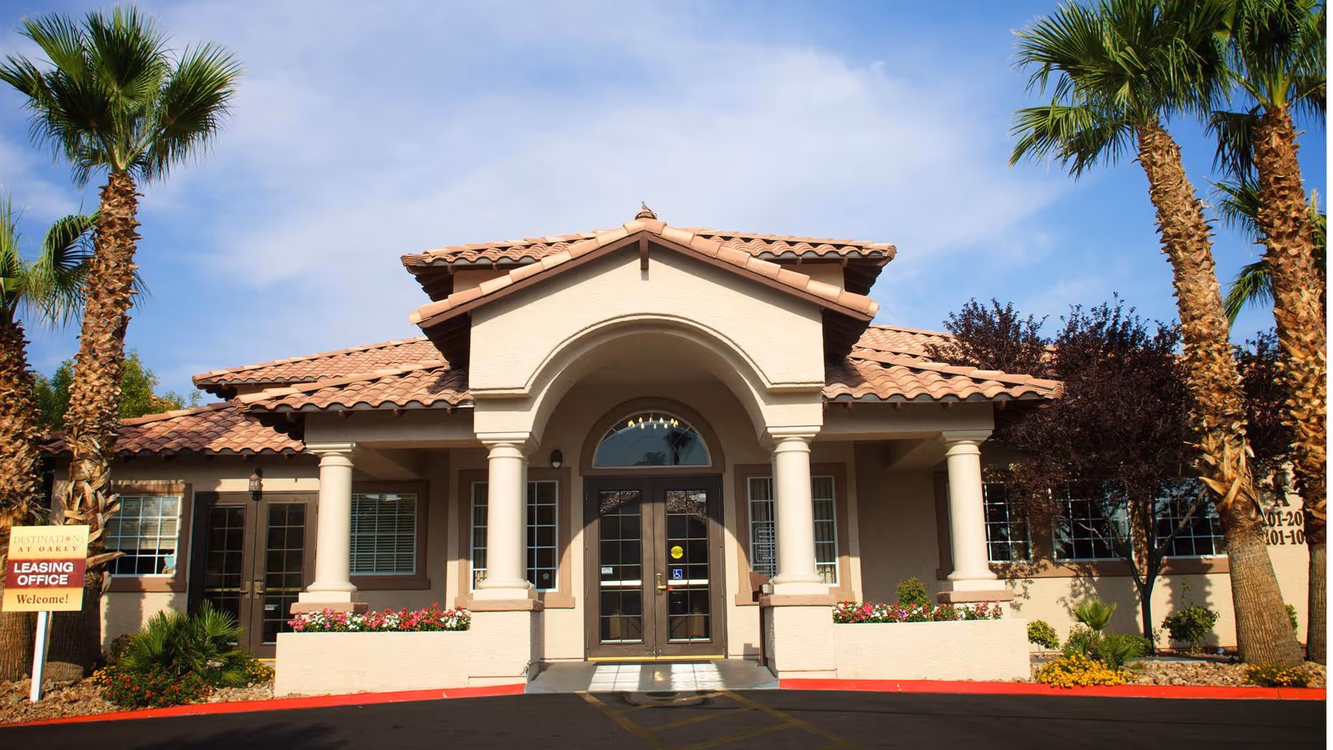 Exterior view of a single-story building with a tiled roof and columns at the entrance, surrounded by palm trees and landscaping. A sign near the entrance reads 'Leasing Office Welcome!'