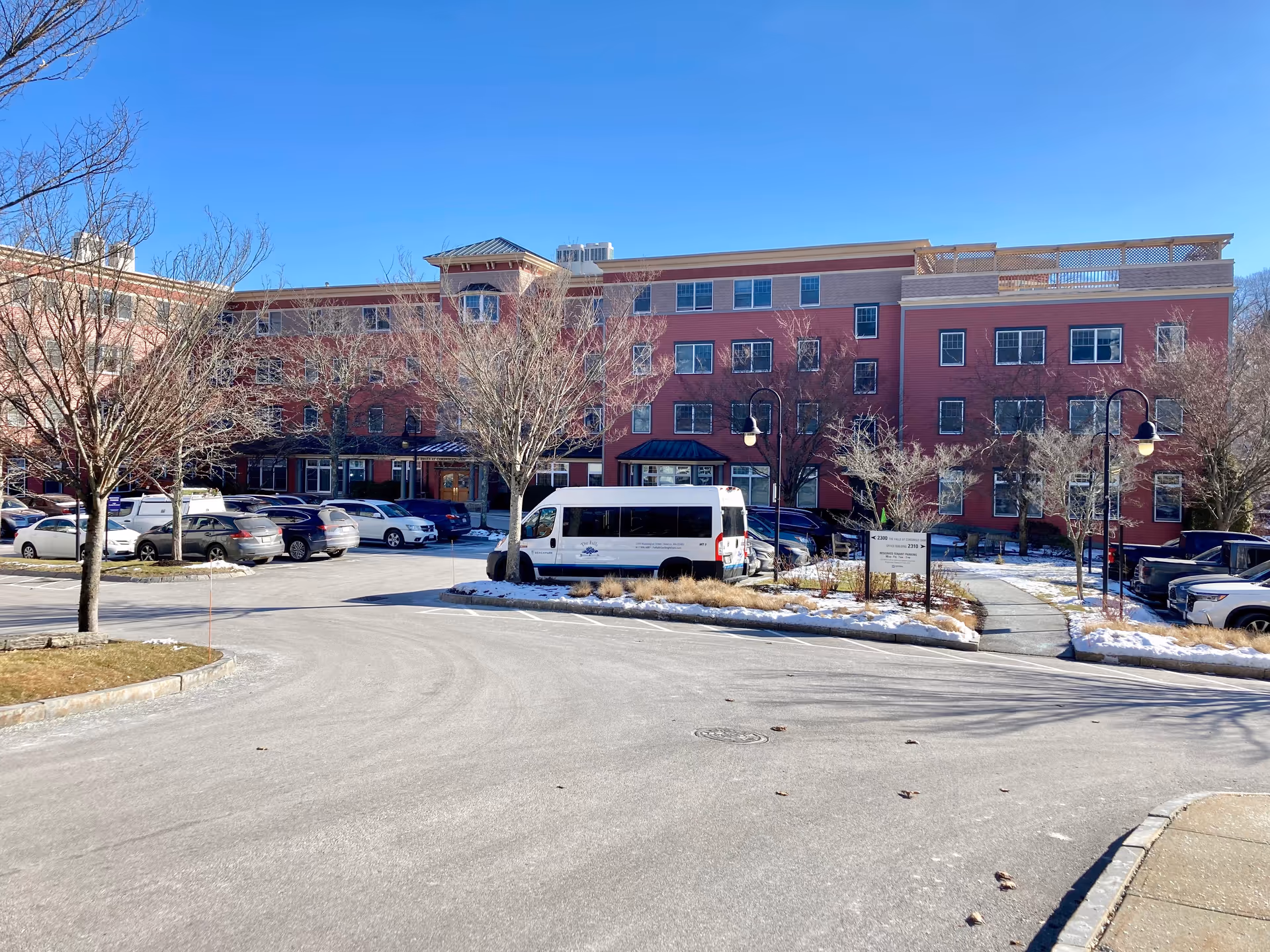 Exterior view of The Falls at Cordingly Dam senior living facility on a clear day, showing a large red brick building with multiple windows, a parking lot with several cars and a white shuttle van, leafless trees, and a pathway with street lamps.