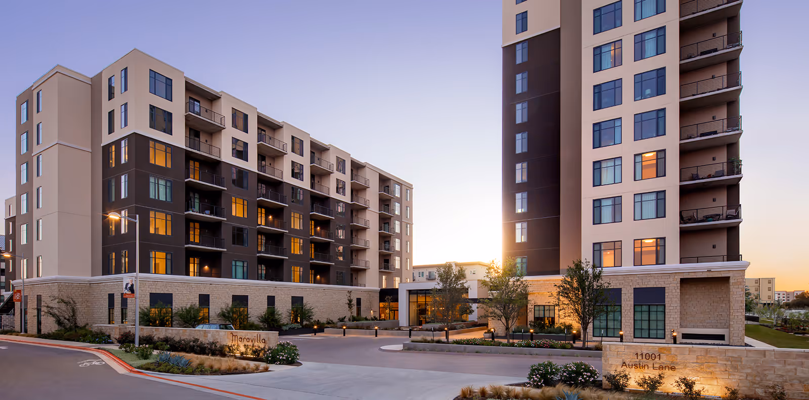 Exterior view of Maravilla at The Domain, a modern multi-story senior living facility with large windows and balconies, surrounded by landscaped greenery and a driveway entrance at sunset.