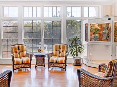 Bright senior living lounge with wicker chairs, a small flower centerpiece, and large windows.