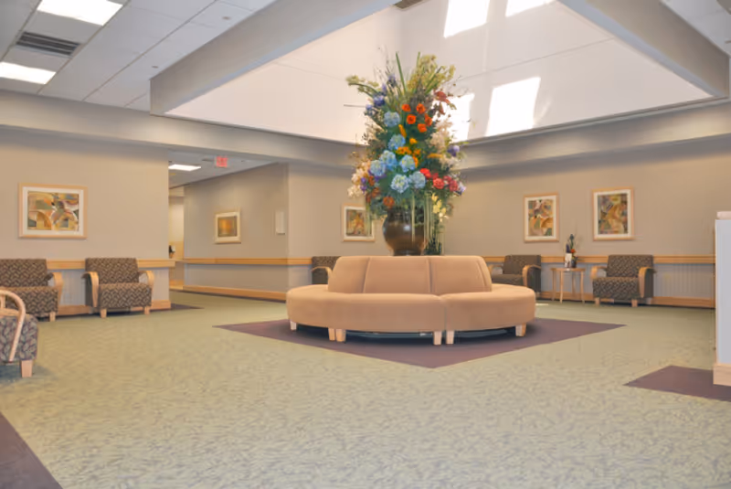 A spacious assisted-living lobby with circular beige seating and a large floral arrangement beneath a skylight.
