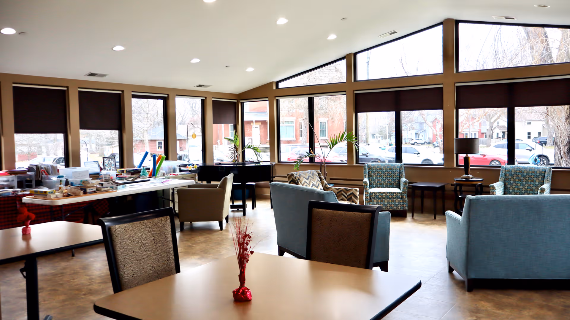 A bright and spacious common area with large windows letting in natural light. The room features several armchairs and sofas arranged for conversation, a piano near the windows, and tables with chairs. One table has a small red decorative centerpiece. There are various items and supplies on a long table along one wall, suggesting a multipurpose or activity space.