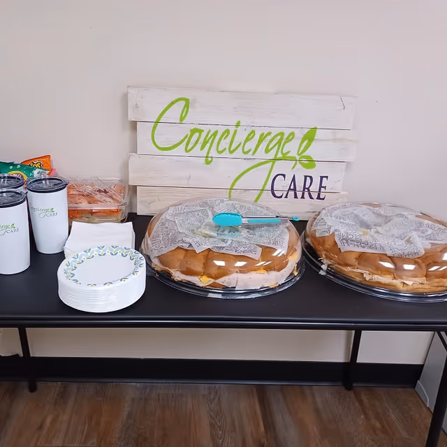 Table with pastries under plastic covers, cups, napkins, and a Concierge Care sign.