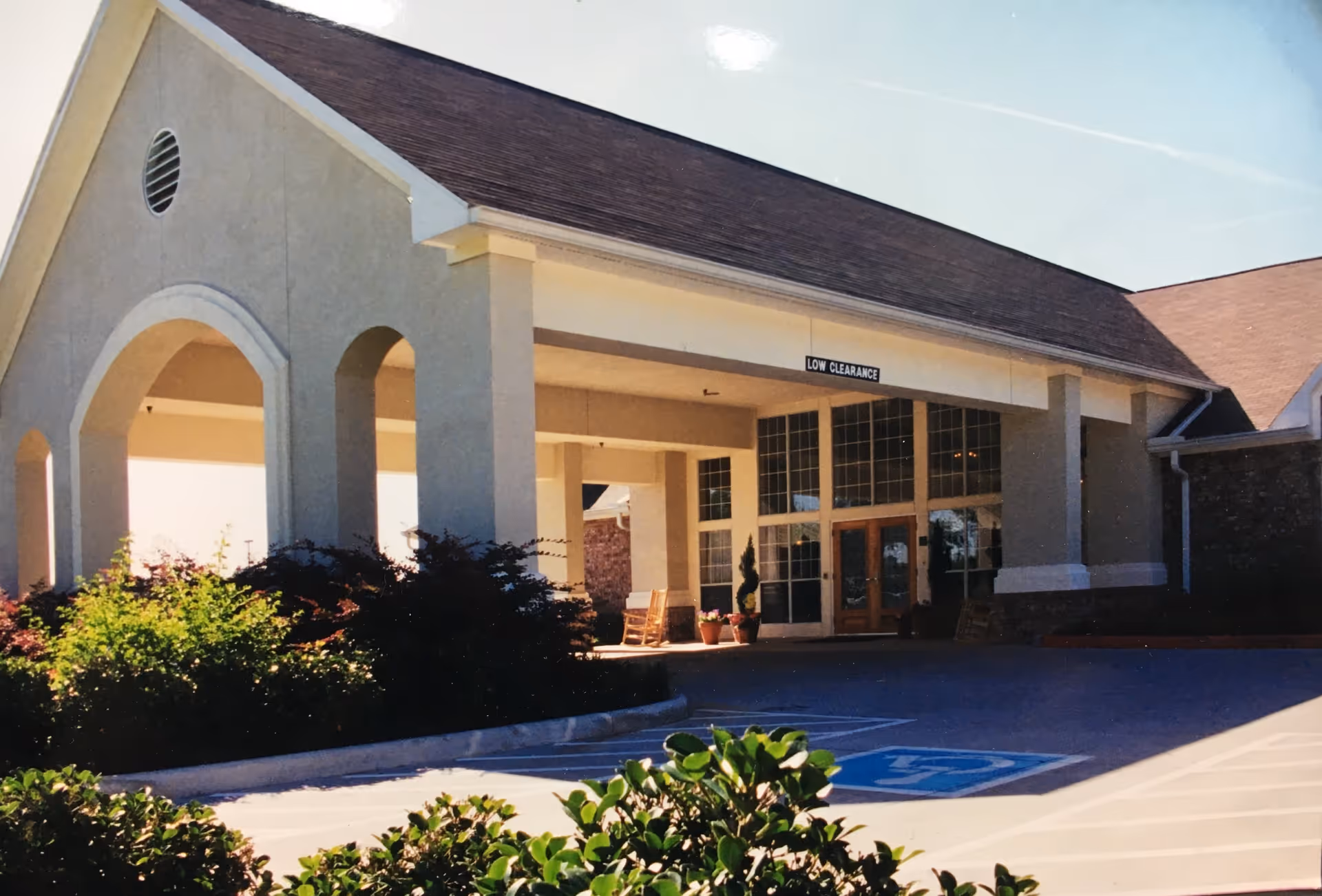 Entrance of a senior living facility with a covered drop-off area, large windows, and a sign indicating low clearance. There are bushes and plants around the driveway and a handicapped parking space visible in front.