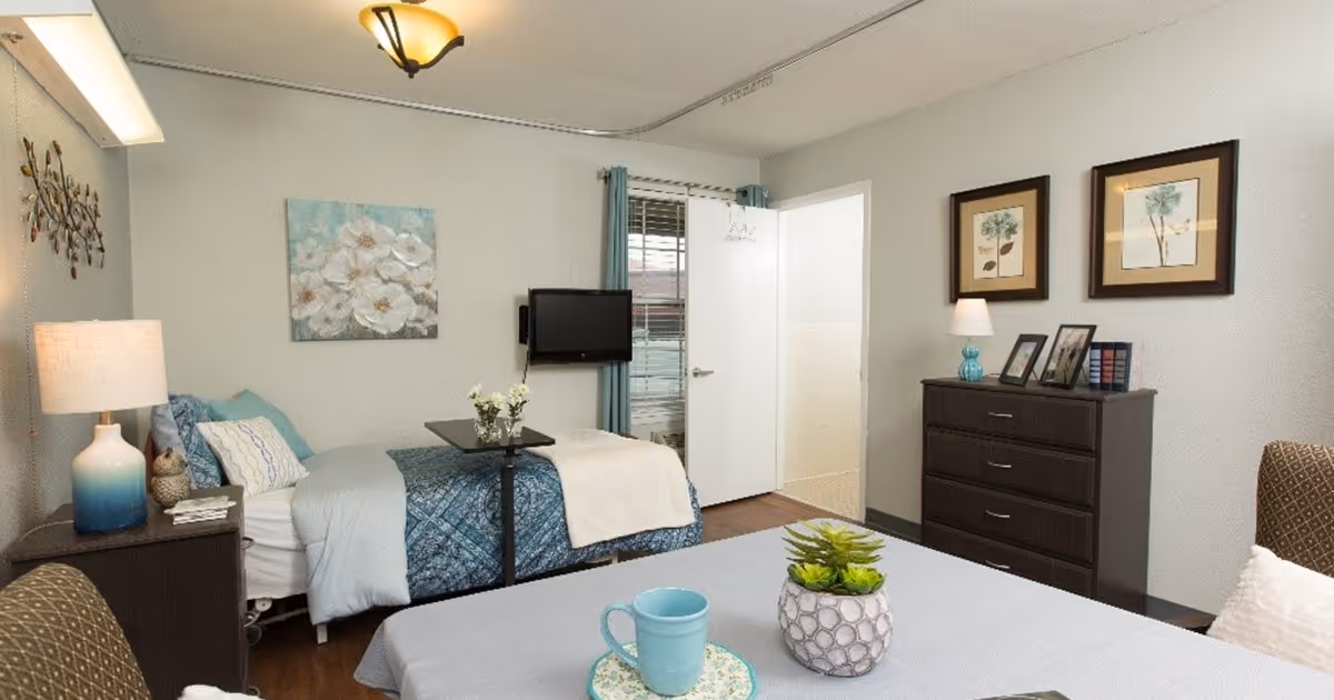 Furnished bedroom with a bed, over-bed tray, TV, dresser, lamps, and decorative artwork.