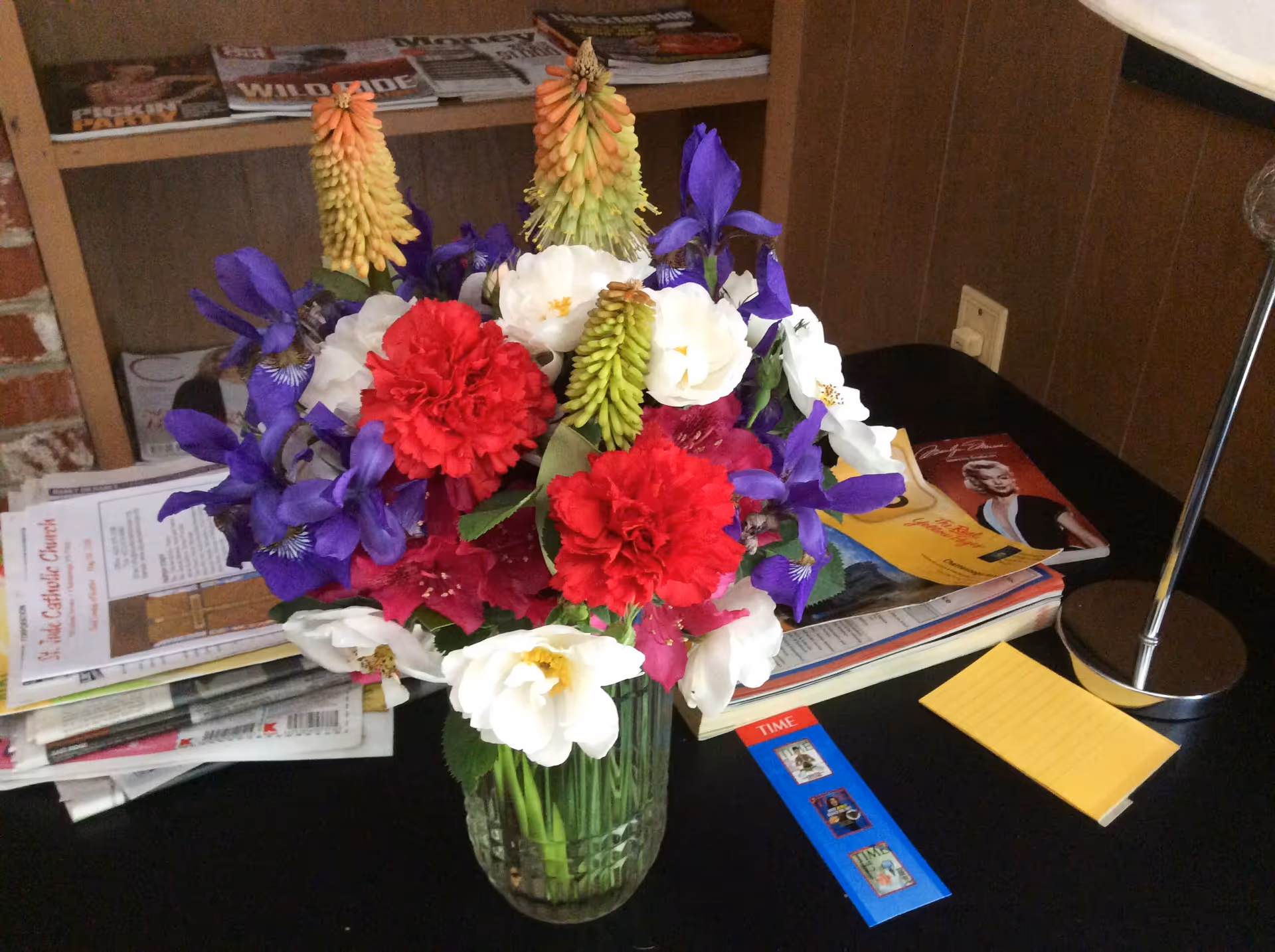 A vase of colorful flowers sits on a table surrounded by magazines, a lamp, and a yellow notepad.