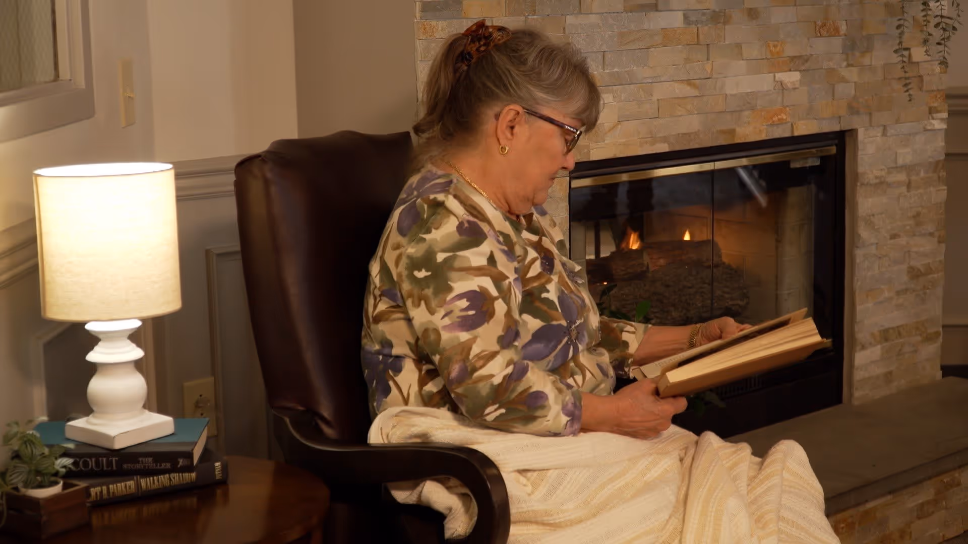 An elderly woman with glasses sits in a dark brown armchair reading a book. She is wrapped in a light-colored blanket and is positioned next to a stone fireplace with a fire burning inside. A small wooden side table with a white lamp, some books, and a small plant is beside her.