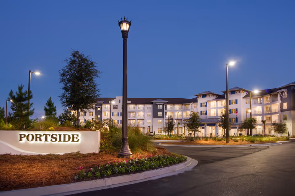 Exterior view of the Portside at Grande Dunes senior living facility at dusk, showing a well-lit multi-story building with balconies, landscaped grounds, street lamps, and a sign that reads 'PORTSIDE'.