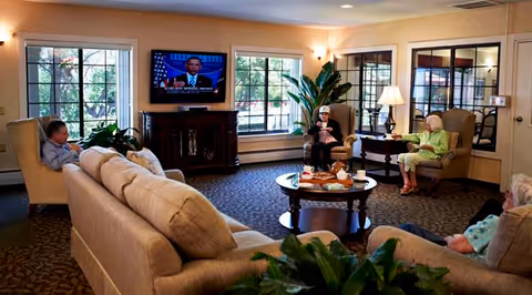 A cozy living room in a senior living facility with beige sofas and armchairs arranged around a round coffee table. Four elderly people are seated, watching a television mounted on the wall displaying a man speaking. Large windows let in natural light, and there are plants and lamps adding to the warm ambiance.
