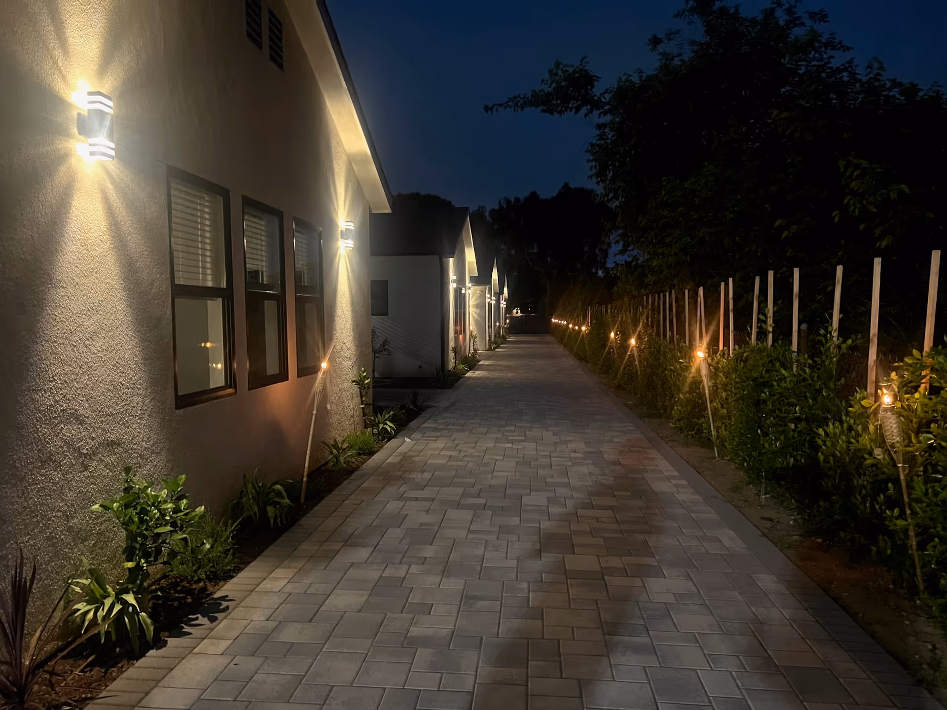 Night view of a lit outdoor walkway beside Ingomar Assisted Living, with wall lights and hedge.