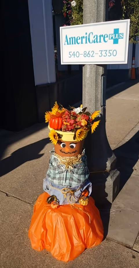 Outdoor scene with AmeriCare Plus sign on a pole and a pumpkin scarecrow decoration.