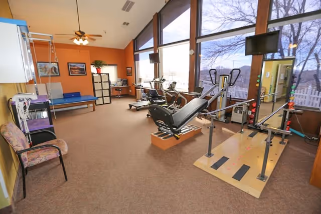 A rehabilitation or physical therapy room with exercise equipment including a leg press machine, parallel bars for walking exercises, and stationary bikes. The room has large windows letting in natural light, a ceiling fan, a chair, and a therapy table against the orange and yellow walls.