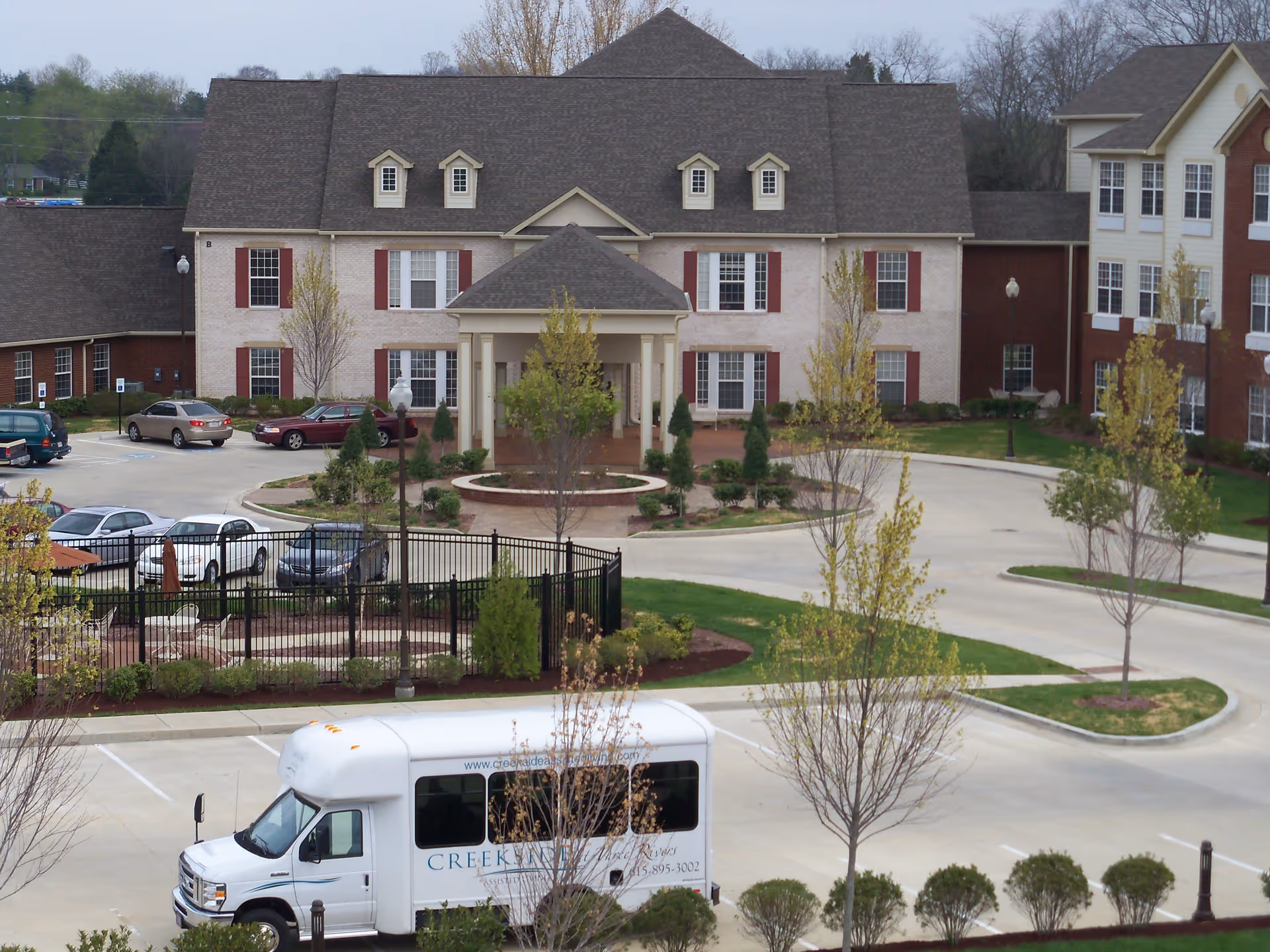 Exterior view of Creekside at Three Rivers Assisted Living facility showing a large building with multiple windows and a covered entrance. There is a circular driveway with landscaped greenery and several parked cars. A white shuttle bus with the facility's name is parked in the foreground.