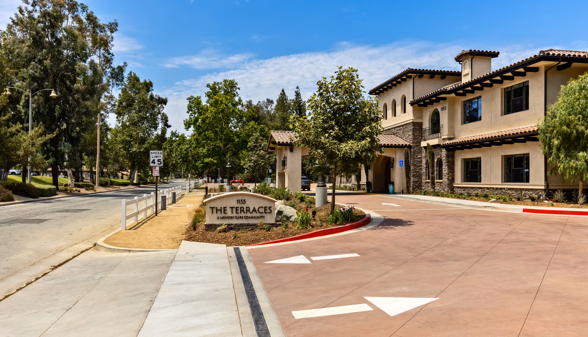 Front entrance and driveway of The Terraces senior living community with a stone sign, landscaped island, and a two-story Mediterranean-style building.
